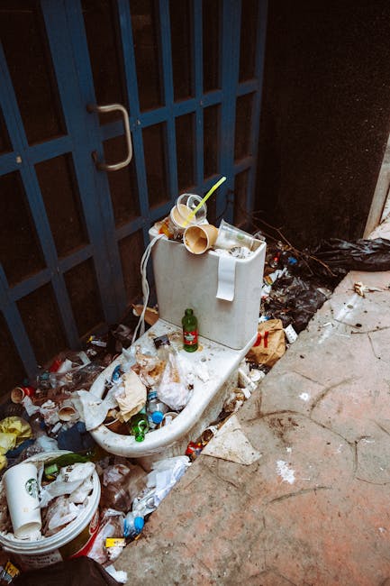 A white ceramic toilet used as a makeshift rubbish disposal area is positioned against a dark blue metal gate on a narrow urban street or alleyway. The toilet is overflowing with various waste materials, including crumpled brown paper, plastic bottles, and discarded food containers, some of which are on top of the toilet tank. Several plastic bottles and cups, along with food packaging, are scattered around the base of the toilet on the ground, contributing to a visibly cluttered and unmanaged waste scene. The surrounding environment features a concrete pavement with cracks and dirt, with a small section of sidewalk visible on the right. A black plastic bag filled with additional rubbish is placed nearby on the ground, and some waste appears to have spilled from it. The overall scene highlights issues related to private waste disposal or alternative rubbish collection methods, such as on-site clearance, managed by waste removal services, with a focus on the visual context of rubbish accumulation in a confined urban setting.