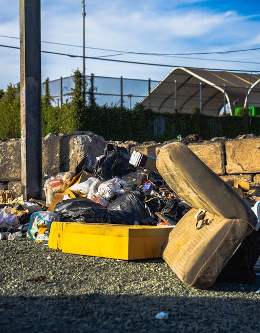 A close-up view of an unmanaged rubbish pile located outdoors, featuring black plastic garbage bags, discarded cardboard, and various waste materials spilling onto a gravel surface. Among the debris, an old, beige car tyre with visible tread and sidewall details is partially resting atop the trash. A bright yellow plastic container lies nearby, contrasting with the dark bags and gravel. In the background, a low stone wall made of irregularly shaped light-colored stones separates the waste area from a fenced tennis court or sports facility, which is partially obscured by green bushes. Metal utility poles and overhead wires are visible against a partly cloudy sky, suggesting an urban or semi-urban location that may be affected by issues of illegal dumping or unauthorized waste discharge. The natural daylight highlights textures and colors, emphasizing the cluttered state that Waste Removal Lewisham might address through alternative waste handling or private rubbish clearance services on narrow urban estates.