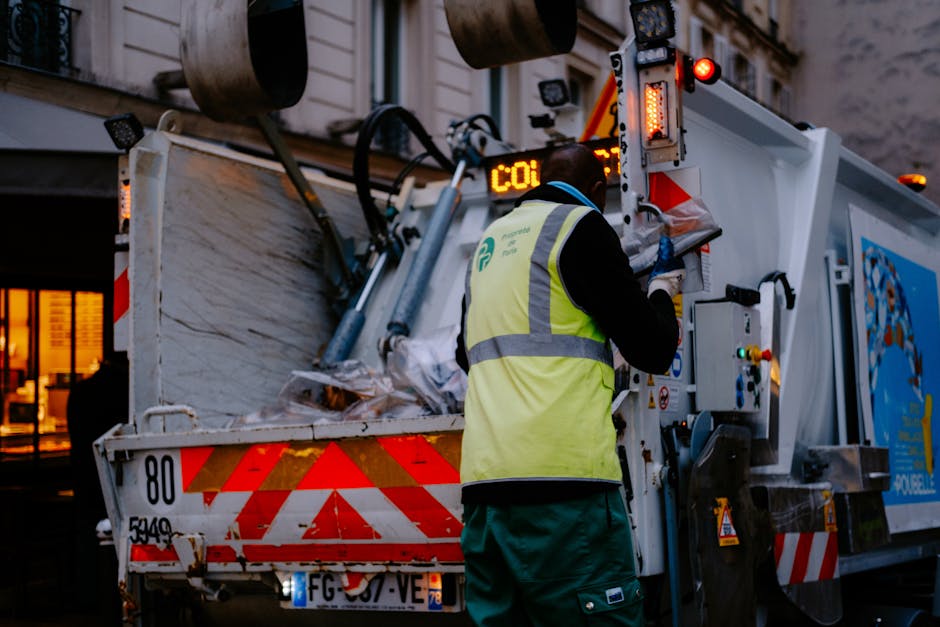 A worker wearing a yellow high-visibility vest and dark clothing operates a waste collection vehicle in an urban setting during evening hours. The worker is standing with their back to the camera, using a control panel on the side of the truck to manage the waste disposal process. The metal body of the waste truck is white with red and orange reflective safety markings, and it contains a partially visible load of mixed waste materials, including paper and plastic bags, inside a large, open-compartment container. The truck's exterior displays safety lights and equipment, with a background of a building with lit windows and street lighting that creates a warm ambient glow. This scene illustrates a private, on-site waste collection service, reflective of independent rubbish removal operations, emphasizing efficient and professional waste handling in an urban environment.