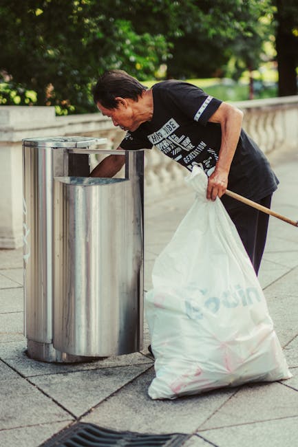 A woman with short dark hair, wearing a black T-shirt with white and blue text and dark trousers, is seen bending over to place a large white plastic bag filled with rubbish into a modern, stainless steel outdoor trash bin. The bin has a cylindrical shape with two side compartments for waste separation, and its surface is smooth and reflective. She is holding the bag with one hand and is using the other hand to steady herself. The scene takes place outdoors on a paved surface, adjacent to a decorative stone balustrade, with lush green foliage and trees in the background, suggesting a park or garden setting. The lighting indicates daytime, with natural light illuminating the scene evenly. This image could relate to the services provided by Waste Removal Lewisham, highlighting practices of on-site waste disposal or independent rubbish collection outside residential or public spaces, illustrating aspects of waste management, disposal, and recycling efforts in Lewisham.