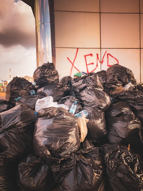 A worker wearing a yellow high-visibility vest and dark clothing operates a waste collection vehicle in an urban setting during evening hours. The worker is standing with their back to the camera, using a control panel on the side of the truck to manage the waste disposal process. The metal body of the waste truck is white with red and orange reflective safety markings, and it contains a partially visible load of mixed waste materials, including paper and plastic bags, inside a large, open-compartment container. The truck's exterior displays safety lights and equipment, with a background of a building with lit windows and street lighting that creates a warm ambient glow. This scene illustrates a private, on-site waste collection service, reflective of independent rubbish removal operations, emphasizing efficient and professional waste handling in an urban environment.