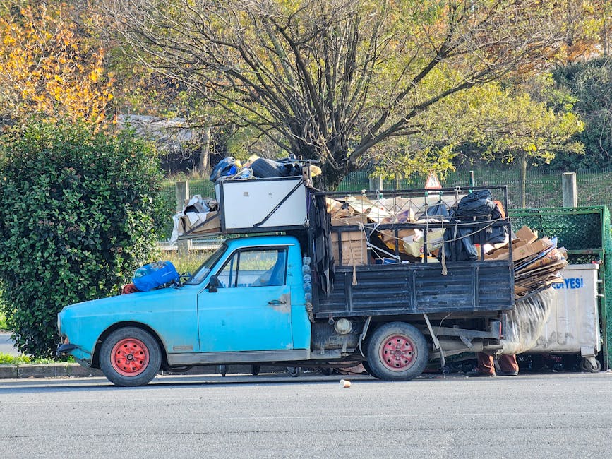 A small blue flatbed truck parked parallel to the pavement with an open cargo area filled with a large volume of mixed waste materials, including flattened cardboard boxes, black plastic rubbish bags, and various debris. The truck's body shows visible signs of wear, with some peeling paint and dirt, and it has red-painted wheels. The cargo is piled high, partially leaning against the truck's metal sides, and appears to be temporarily stored before disposal or removal. Behind the truck, there is a row of green metal fencing, with a backdrop of trees and shrubs displaying autumn foliage, including yellow and green leaves, and sunlight filtering through the branches. The scene suggests a private waste collection or on-site clearance operation, supported by the presence of waste removal services that handle bulky rubbish not suitable for regular municipal collection.