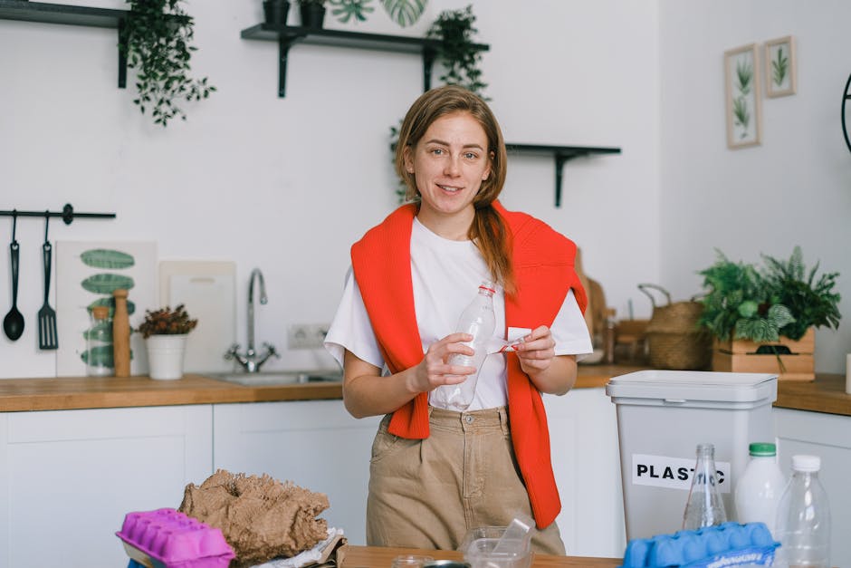 A young woman with light skin, shoulder-length light brown hair, and a friendly expression, is standing in a well-lit modern kitchen. She is wearing a white t-shirt and light beige trousers, with a bright orange sweater draped over her shoulders. She is holding a clear plastic water bottle in her right hand and a small packet or wrapper in her left hand. The background features a wooden countertop with various small potted plants, kitchen utensils hanging on a black rail, and a white wall with framed botanical prints. To her right, there is a white plastic bin labeled 'PLASTIC' for waste separation, alongside a few plastic bottles and containers. The scene appears to depict a domestic setting focused on recycling or waste sorting, aligning with the concept of alternative waste handling or rubbish management, as typical of services offered by Waste Removal Lewisham. The overall atmosphere is clean and organized, emphasizing the importance of proper rubbish disposal within a household environment.