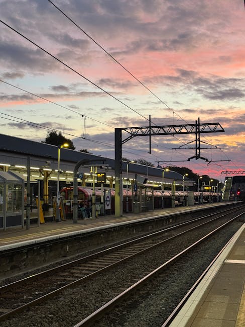 A view of Lewisham railway station platform during dusk, with multiple train carriages parked alongside the shelter. The platform is illuminated by yellowish lights, casting a warm glow over the waiting area. Overhead, there are electrical wires supported by metal poles and structures, with a sky painted in pink, purple, and orange hues from the setting sun. The scene shows the tracks running parallel to the platform, with a clean, paved edge and safety barriers. The environment is calm and quiet, typical of early evening, and the image reflects a typical urban railway setting suitable for situational context related to alternative waste handling or private rubbish collection at transportation hubs, with Lewisham's train station backdrop highlighting commuter infrastructure.