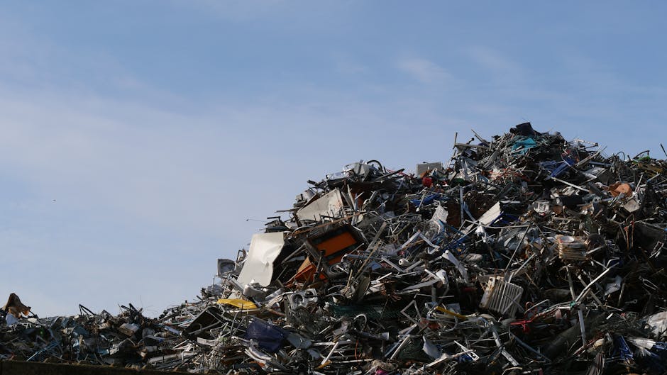 A large mound of mixed waste materials piled outdoors under a clear blue sky, consisting of various types of metal, plastic, and electronic debris. The heap includes disorganized scrap appliances, framework, and smaller discarded items, with some parts appearing twisted or broken. The materials show a range of textures from smooth plastic surfaces to rough, jagged metal edges, with colors primarily metallic silver, black, and occasional hints of orange, blue, and green. The waste is densely packed and loosely stacked, occupying most of the foreground with no clear boundaries, while the background features an open sky. The scene reflects a site of significant rubbish accumulation, likely from a mechanical or waste disposal process, emphasizing the importance of controlled waste removal services for proper management and environmental protection. Waste Removal Lewisham occasionally handles such bulky, mixed debris as part of alternative waste handling solutions to prevent illegal dumping and promote responsible disposal practices.