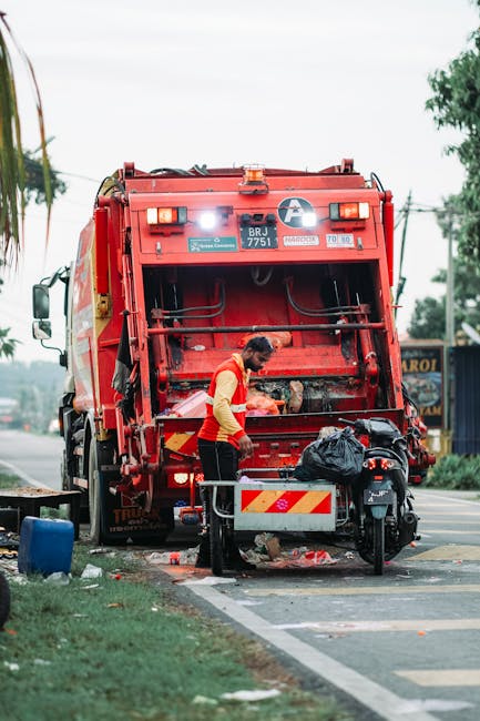 A waste collection worker wearing a high-visibility orange and yellow vest is operating a red refuse collection vehicle parked on the side of a street. The back of the vehicle is open, exposing mechanical parts and storage compartments, with the worker positioned in front of the loading area, handling black plastic rubbish bags placed on a small metal platform attached to the vehicle. The street scene includes a paved sidewalk, a grassy verge, and in the background, there are trees, utility poles, and building facades, indicating an urban or suburban environment. The lighting appears natural and overcast, with diffused daylight illuminating the scene. This setting reflects an independent waste collection process, highlighting how private rubbish removal services like Waste Removal Lewisham manage refuse collection outside of municipal systems, capturing the practical activity involved in efficient rubbish disposal and on-site clearance.