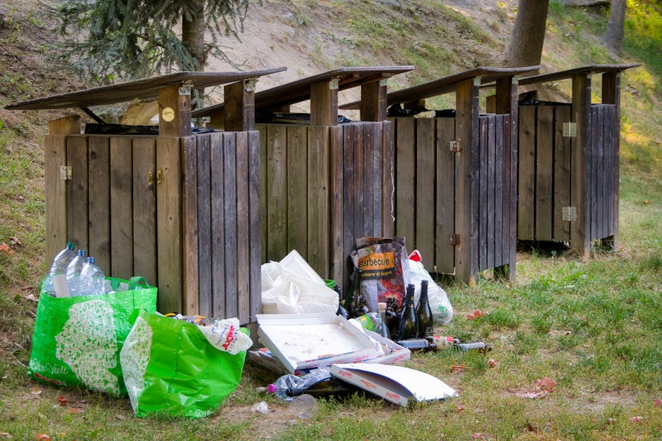A large, densely packed pile of mixed waste materials occupying an outdoor area, consisting predominantly of plastic waste such as crumpled and torn plastic bags, wrappers, and bottles, along with paper, cardboard, and some organic matter. The plastics vary in color, including red, white, blue, and transparent, with some items partially buried beneath others. Smaller debris such as food packaging, bottles, and miscellaneous refuse are scattered across the mound. In the center of the image, there is a worn, green plastic bucket nestled among the rubbish, indicating possible informal collection or disposal. Surrounding the pile, the environment appears to be an open, unpaved area, with a slight slope and uneven ground surface partially visible at the edges. The lighting suggests natural daylight with shadows cast across different waste objects, emphasizing the chaotic and unmanaged state of the debris. This scene exemplifies the type of unmanaged waste accumulation that private waste removal services like Waste Removal Lewisham often address through on-site clearance or independent collection solutions, highlighting the importance of effective rubbish management in preventing environmental pollution and maintaining public health in residential or commercial areas.