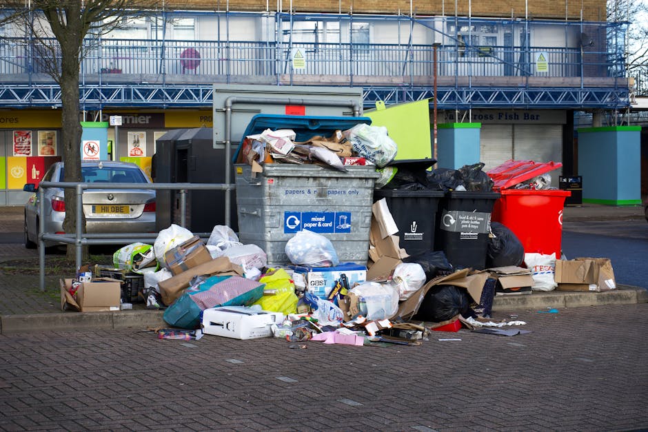 In the foreground of the image, multiple overflowing rubbish bins and waste collection containers are seen on a paved sidewalk area, with various types of waste spilling out and scattered around. The central bin is a large, grey, multi-purpose container made of plastic, designated for mixed paper and card, filled with crumpled newspapers, cardboard boxes, and paper packaging. To its right are two black bins, one with a lid slightly ajar and filled with general waste, and a red bin behind them. Additional waste, including flattened cardboard boxes, plastic bags, and smaller packaging items, are piled on the ground around the bins, some leaning against the containers and others strewn across the pavement. In the background, a parked silver car is partially visible behind a metal railing, with a row of shopfronts featuring signs and storefronts further behind. Behind these, a blue scaffolding structure surrounds a building under construction or renovation, with a commercial property sign partially visible. The scene suggests an instance of private waste accumulation, highlighting the need for professional rubbish removal services.