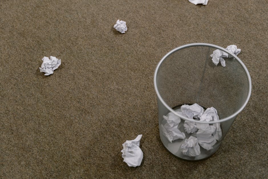 The image depicts a transparent cylindrical waste bin made of plastic sitting on a brown carpeted floor. The bin contains crumpled white paper, with several additional crumpled sheets scattered on the surrounding area of the carpet, indicating a scene of waste disposal or tidying up. The paper pieces are irregular in shape, with visible creases and folds, and vary in size. The lighting appears natural and even, highlighting the texture of the carpet and the crumpled paper without strong shadows. The environment suggests an indoor setting, possibly an office or home workspace, with the focus on waste collection and rubbish disposal. This visual aligns with private rubbish removal services provided by Waste Removal Lewisham, highlighting the need for efficient on-site clearance of paper waste as part of general rubbish management tasks.