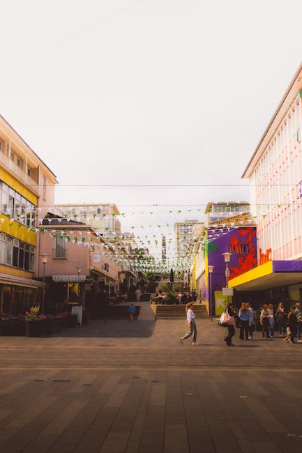 In the foreground of the image, multiple overflowing rubbish bins and waste collection containers are seen on a paved sidewalk area, with various types of waste spilling out and scattered around. The central bin is a large, grey, multi-purpose container made of plastic, designated for mixed paper and card, filled with crumpled newspapers, cardboard boxes, and paper packaging. To its right are two black bins, one with a lid slightly ajar and filled with general waste, and a red bin behind them. Additional waste, including flattened cardboard boxes, plastic bags, and smaller packaging items, are piled on the ground around the bins, some leaning against the containers and others strewn across the pavement. In the background, a parked silver car is partially visible behind a metal railing, with a row of shopfronts featuring signs and storefronts further behind. Behind these, a blue scaffolding structure surrounds a building under construction or renovation, with a commercial property sign partially visible. The scene suggests an instance of private waste accumulation, highlighting the need for professional rubbish removal services.